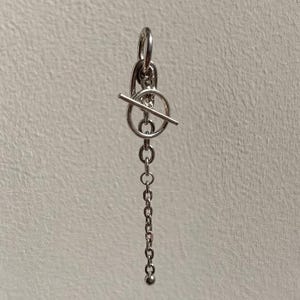 May include: A silver-coloured metal earring featuring a toggle clasp mechanism with a circular ring and a horizontal bar. A delicate chain hangs downwards from the toggle, ending in a small, smooth spherical bead at the bottom of the dangling chain.