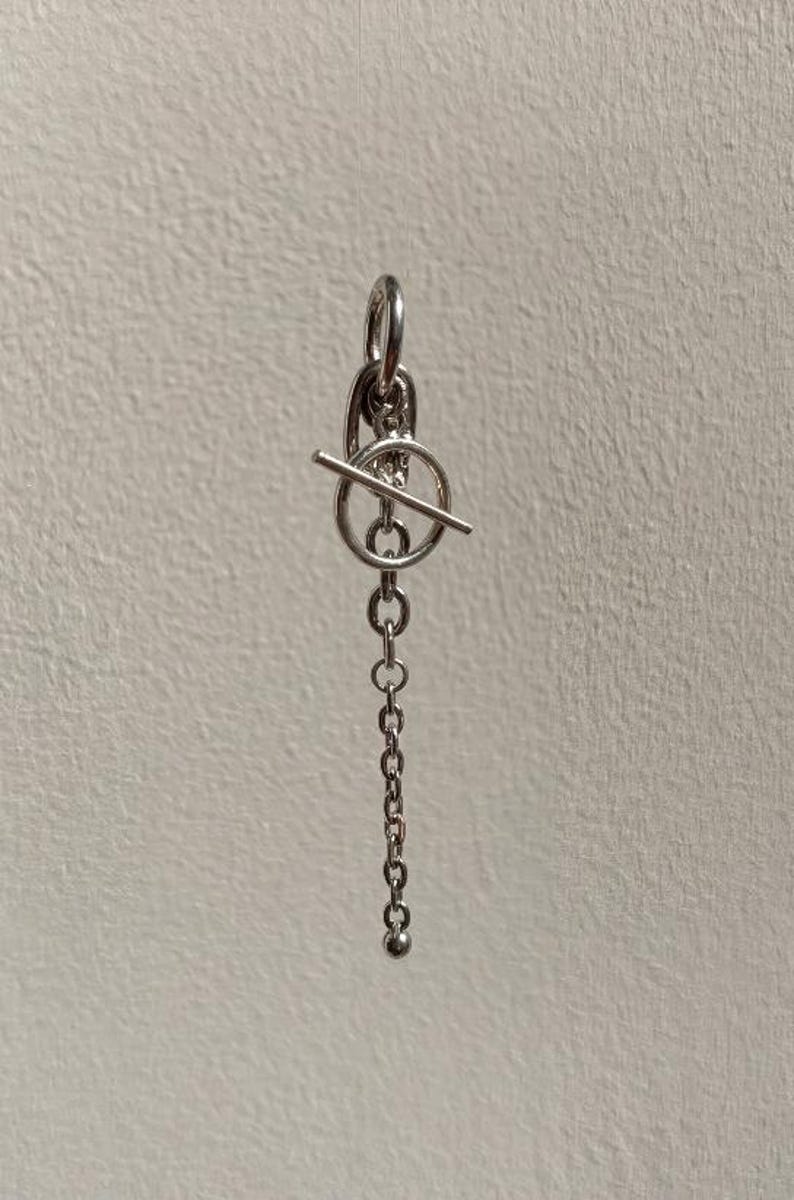 May include: A silver-coloured metal earring featuring a toggle clasp mechanism with a circular ring and a horizontal bar. A delicate chain hangs downwards from the toggle, ending in a small, smooth spherical bead at the bottom of the dangling chain.