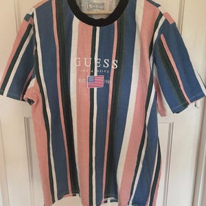 Peut inclure: Un t-shirt à manches courtes avec des rayures verticales roses, bleues, blanches et vert foncé. Le vêtement présente un col rond noir et affiche le texte GUESS LOS ANGELES EST 1981 avec un petit écusson drapeau américain sur la poitrine.
