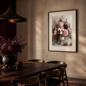 May include: A framed wall art print depicting a floral arrangement in a pedestal vase, featuring shades of pink, burgundy, and cream flowers with green foliage. The artwork is displayed in a dark frame on a textured beige wall above a wooden table.