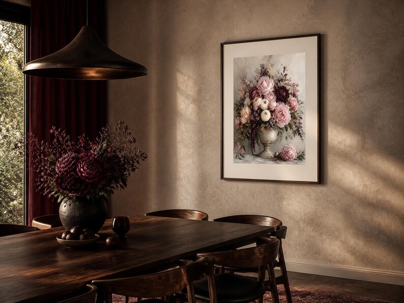 May include: A framed wall art print depicting a floral arrangement in a pedestal vase, featuring shades of pink, burgundy, and cream flowers with green foliage. The artwork is displayed in a dark frame on a textured beige wall above a wooden table.