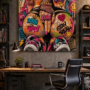 Op de afbeelding: Een vierkant wandkunstwerk met levendige graffiti-stijl straatkunst. De centrale afbeelding toont een persoon met visnet-handschoenen en sneakers, omringd door kleurrijke tags en teksten zoals BORN TO WIN, LOVE en STAY REAL.