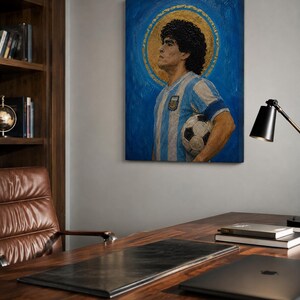 May include: A rectangular canvas painting depicting a man with dark curly hair wearing a light blue and white striped jersey, holding a soccer ball. A gold circular halo surrounds the head against a textured blue background, creating a portrait.