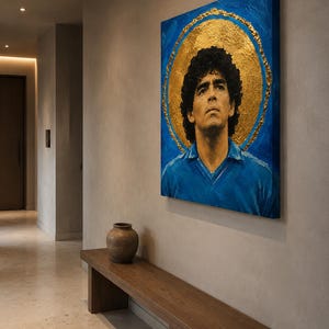 Puede incluir: Un cuadro cuadrado sobre lienzo que representa un retrato de Diego Maradona con una camiseta azul, sobre un fondo azul vibrante con un gran halo circular de pan de oro texturizado que rodea la cabeza, montado en una pared de tonos neutros.