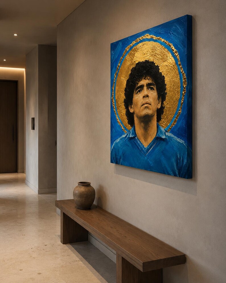 Puede incluir: Un cuadro cuadrado sobre lienzo que representa un retrato de Diego Maradona con una camiseta azul, sobre un fondo azul vibrante con un gran halo circular de pan de oro texturizado que rodea la cabeza, montado en una pared de tonos neutros.