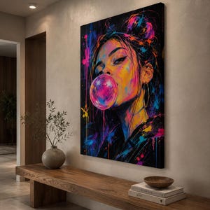 Puede incluir: Un lienzo rectangular para pared que muestra un retrato vibrante y colorido de una persona con el cabello recogido en un mo&ntilde;o, soplando una gran burbuja de chicle rosa. La obra utiliza salpicaduras de pintura ne&oacute;n rosa, azul y amarilla.