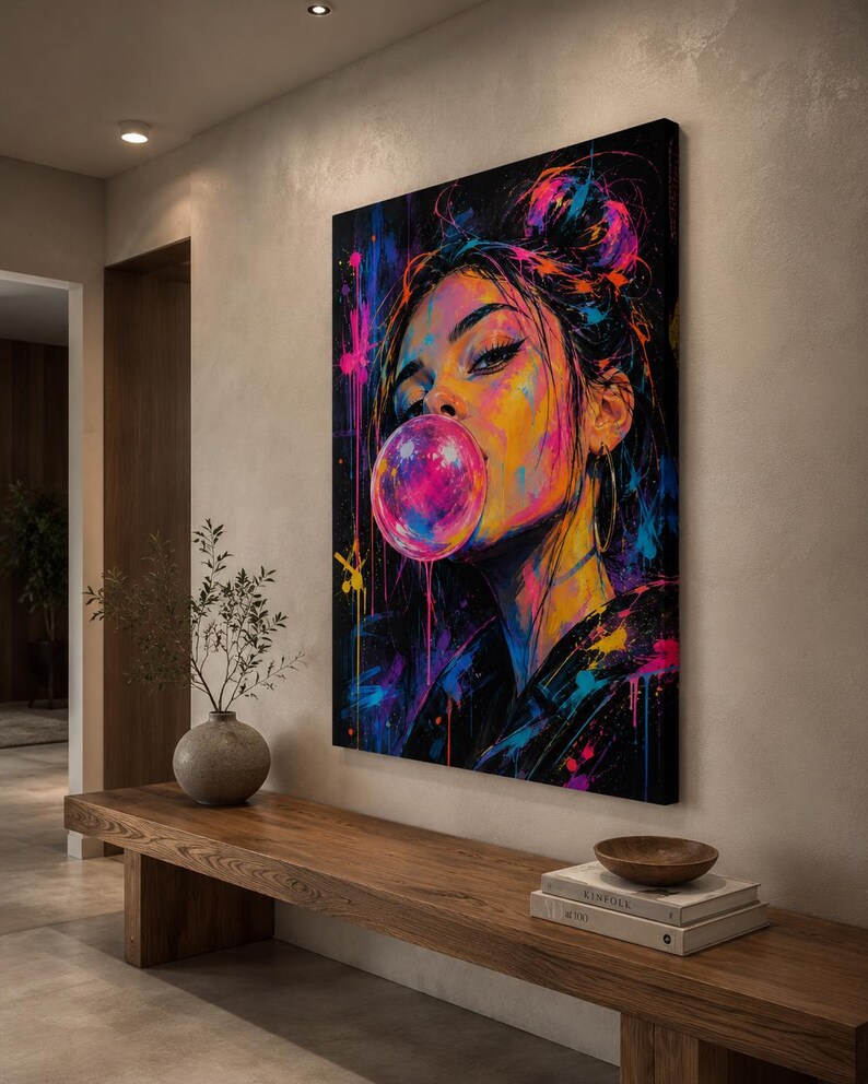 Puede incluir: Un lienzo rectangular para pared que muestra un retrato vibrante y colorido de una persona con el cabello recogido en un mo&ntilde;o, soplando una gran burbuja de chicle rosa. La obra utiliza salpicaduras de pintura ne&oacute;n rosa, azul y amarilla.