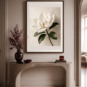 Puede incluir: Una l&aacute;mina de arte mural enmarcada que muestra una flor de magnolia blanca con hojas de color verde oscuro sobre un fondo beige claro. La obra se exhibe sobre una mesa consola beige curva con un jarr&oacute;n oscuro y un cuenco peque&ntilde;o.