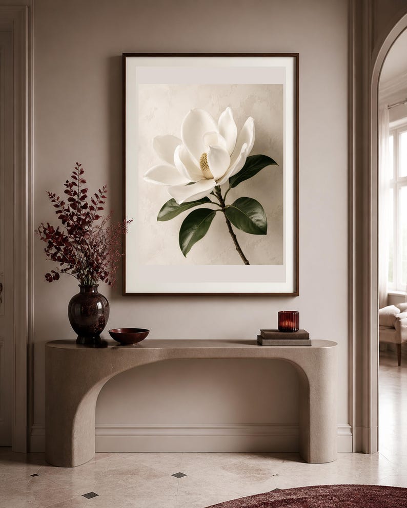 Puede incluir: Una l&aacute;mina de arte mural enmarcada que muestra una flor de magnolia blanca con hojas de color verde oscuro sobre un fondo beige claro. La obra se exhibe sobre una mesa consola beige curva con un jarr&oacute;n oscuro y un cuenco peque&ntilde;o.