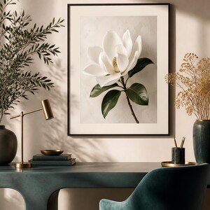 Puede incluir: Una impresi&oacute;n de arte mural enmarcada que presenta una sola flor de magnolia blanca con hojas de color verde oscuro sobre un fondo claro y texturizado. La obra de arte se exhibe en un marco negro delgado, mostrando el tema bot&aacute;nico con p&eacute;talos claros y detallados.