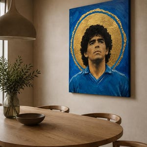 Puede incluir: Un cuadro rectangular sobre lienzo que muestra el retrato de un hombre de pelo oscuro y rizado con una camisa de cuello azul. La figura est&aacute; centrada sobre un fondo azul vibrante con un gran halo circular de pan de oro texturizado detr&aacute;s.