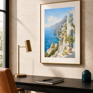 May include: A framed wall art print depicting a coastal Mediterranean scene with white buildings, a golden domed church, a blue sea, and a potted lemon tree in the foreground. The artwork is displayed in a thin gold frame on a textured wall.