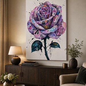 Puede incluir: Un cuadro rectangular sobre lienzo que presenta una gran rosa morada central cubierta de coloridos grafitis, s&iacute;mbolos y texto. La rosa tiene un tallo verde con dos hojas, todo ello acentuado por salpicaduras de pintura negra sobre un fondo blanco.