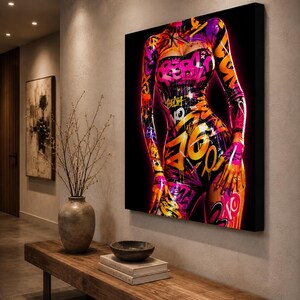Puede incluir: Un lienzo rectangular para pared que presenta una vibrante ilustraci&oacute;n estilo grafiti de un torso. La obra muestra patrones de pintura en aerosol rosa ne&oacute;n, naranja y morado, incluyendo la palabra REBEL, sobre un fondo negro profundo.