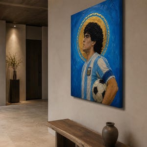 May include: A square canvas painting featuring a portrait of a soccer player with dark curly hair, wearing a light blue and white striped jersey with an emblem, holding a soccer ball, set against a vibrant blue background with a gold halo.