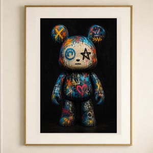 Op de afbeelding: Een ingelijste wandkunstprint met een gestileerde berenfiguur bedekt met levendige graffiti-achtige verfspatten, waaronder een blauwe cirkel, een zwarte ster en een rood hart, tegen een donkere achtergrond in een dunne goudkleurige lijst.