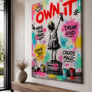 May include: A rectangular canvas wall art piece featuring a black and white illustration of a child wearing a crown, standing on a wooden crate and spray painting the text OWN IT in red. The background is a vibrant splatter of neon paint colors.