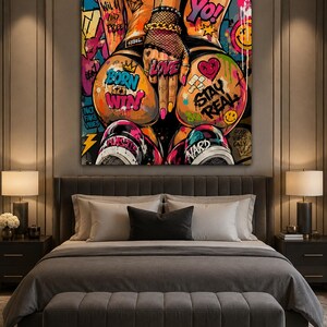 Op de afbeelding: Een vierkant kunstwerk voor aan de muur in een levendige graffitistijl. De afbeelding toont de onderrug en heupen van een persoon, versierd met kleurrijke tatoeages en teksten zoals BORN TO WIN, STAY REAL, LOVE en YO tegen een chaotische achtergrond.
