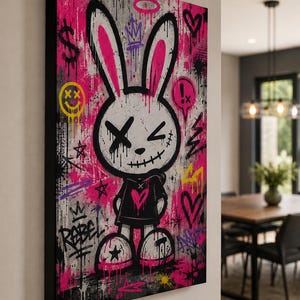 May include: A rectangular canvas wall art piece featuring a stylized white rabbit character with black stitched eyes and mouth, wearing a black hoodie with a pink heart. The background is a vibrant mix of pink and gray graffiti with symbols.