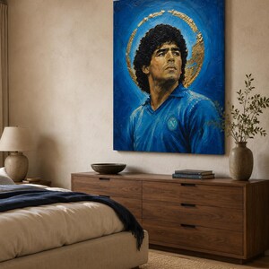 Puede incluir: Un cuadro rectangular sobre lienzo que muestra el retrato de un hombre de pelo oscuro y rizado con una camisa azul de cuello con un logotipo circular en el pecho. Un efecto de halo dorado texturizado rodea la cabeza sobre un fondo azul intenso.