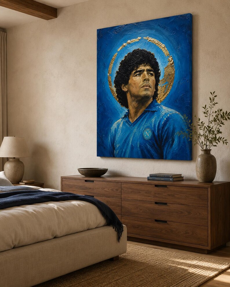Puede incluir: Un cuadro rectangular sobre lienzo que muestra el retrato de un hombre de pelo oscuro y rizado con una camisa azul de cuello con un logotipo circular en el pecho. Un efecto de halo dorado texturizado rodea la cabeza sobre un fondo azul intenso.