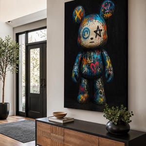 Op de afbeelding: Een groot rechthoekig wandkunstwerk met een gestileerde berenfiguur tegen een effen zwarte achtergrond. De beer is bedekt met levendige graffiti-achtige verfspatten, waaronder blauw, oranje en rood, met symbolen zoals een hart en een ster.