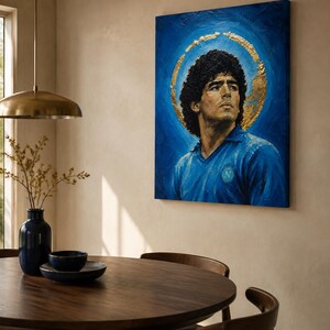 Puede incluir: Un cuadro rectangular sobre lienzo que muestra el retrato de un hombre de pelo oscuro y rizado con una camiseta azul y un emblema circular. El fondo es de color azul intenso con un halo de pan de oro texturizado alrededor de la cabeza.