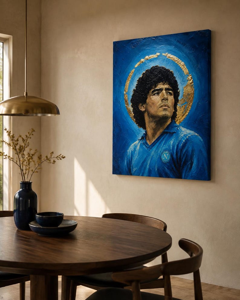 Puede incluir: Un cuadro rectangular sobre lienzo que muestra el retrato de un hombre de pelo oscuro y rizado con una camiseta azul y un emblema circular. El fondo es de color azul intenso con un halo de pan de oro texturizado alrededor de la cabeza.