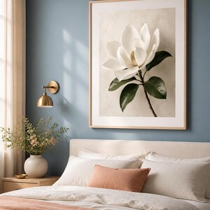 Puede incluir: Una l&aacute;mina de arte enmarcada que muestra una gran flor de magnolia blanca con hojas de color verde oscuro sobre un fondo beige texturizado. La obra est&aacute; expuesta en un marco de madera clara y cuelga en una pared azul sobre una cama.