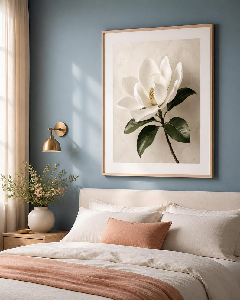 Puede incluir: Una l&aacute;mina de arte enmarcada que muestra una gran flor de magnolia blanca con hojas de color verde oscuro sobre un fondo beige texturizado. La obra est&aacute; expuesta en un marco de madera clara y cuelga en una pared azul sobre una cama.