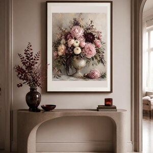 May include: A framed wall art print featuring a floral arrangement in a pedestal vase, displaying pink, white, and deep burgundy flowers with green foliage. The artwork is displayed above a curved console table with a dark vase and a small bowl.