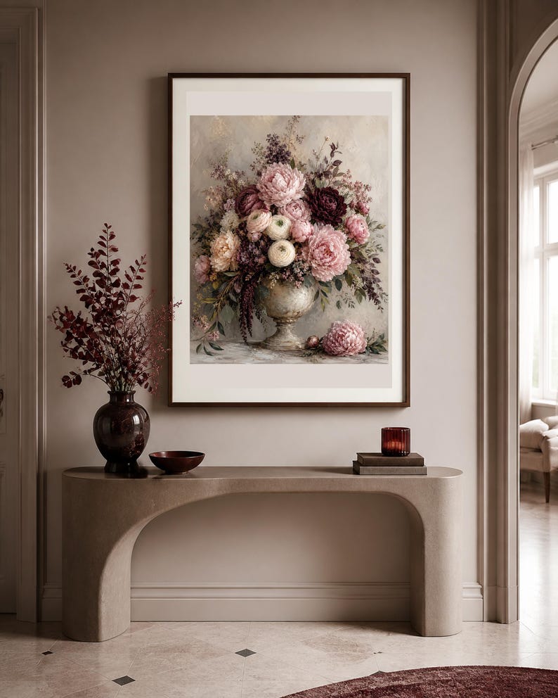 May include: A framed wall art print featuring a floral arrangement in a pedestal vase, displaying pink, white, and deep burgundy flowers with green foliage. The artwork is displayed above a curved console table with a dark vase and a small bowl.