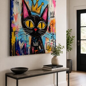 Pode incluir: Uma tela quadrada apresentando um gato preto estilizado com grandes olhos amarelos e uma coroa amarela. O fundo &eacute; uma mistura vibrante de respingos de tinta azul, rosa e amarela, marcas estilo grafite, cora&ccedil;&otilde;es e s&iacute;mbolos abstratos.