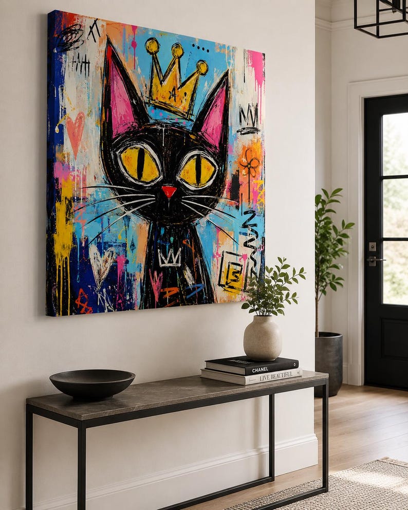 Pode incluir: Uma tela quadrada apresentando um gato preto estilizado com grandes olhos amarelos e uma coroa amarela. O fundo &eacute; uma mistura vibrante de respingos de tinta azul, rosa e amarela, marcas estilo grafite, cora&ccedil;&otilde;es e s&iacute;mbolos abstratos.