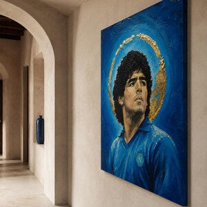 Puede incluir: Un cuadro rectangular sobre lienzo que muestra un retrato de Diego Maradona con una camiseta azul y un halo circular de pan de oro sobre un fondo azul intenso. La obra est&aacute; expuesta en una pared clara en un pasillo con una entrada arqueada.