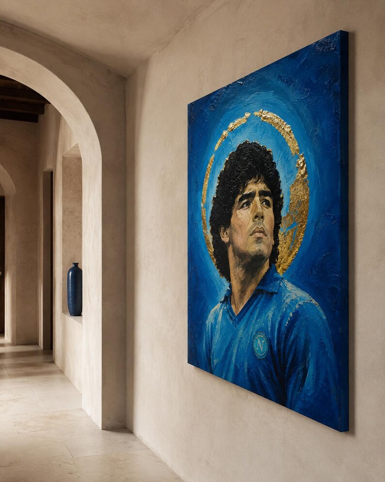 Puede incluir: Un cuadro rectangular sobre lienzo que muestra un retrato de Diego Maradona con una camiseta azul y un halo circular de pan de oro sobre un fondo azul intenso. La obra est&aacute; expuesta en una pared clara en un pasillo con una entrada arqueada.
