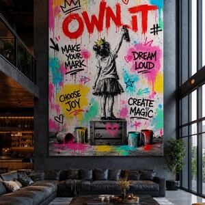 May include: A large rectangular wall art piece featuring a child wearing a crown, standing on a wooden crate and spray painting the text "OWN IT" in red. The background is a vibrant mix of colorful paint splatters, hearts, and stars with the phrases "MAKE YOUR MARK," "DREAM LOUD," "CHOOSE JOY," and "CREATE MAGIC."