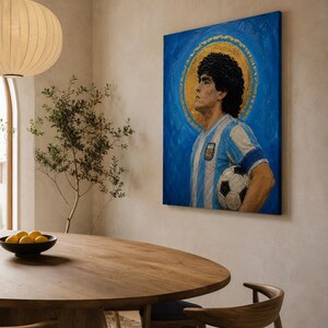 May include: A rectangular canvas painting featuring a portrait of a soccer player with dark curly hair, wearing a light blue and white striped jersey, holding a soccer ball, set against a vibrant blue background with a circular gold halo.