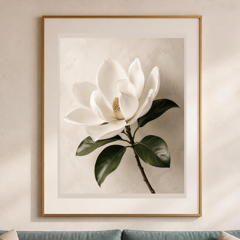 Puede incluir: Una l&aacute;mina de arte enmarcada que muestra una gran flor de magnolia blanca con centro amarillo y hojas de color verde oscuro. El motivo bot&aacute;nico est&aacute; centrado sobre un fondo beige neutro y texturizado, dentro de un marco met&aacute;lico dorado.