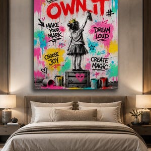 May include: A rectangular wall art print featuring a child wearing a crown standing on a wooden crate, spray painting the text OWN IT. The background is a vibrant graffiti-style splash of pink, yellow, and blue with the phrases MAKE YOUR MARK, DREAM LOUD, CHOOSE JOY, and CREATE MAGIC.