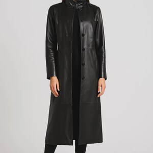 May include: A long black leather coat featuring a high stand-up collar, long sleeves, and a front button closure. The coat extends to the mid-calf and is worn over a black top, black trousers, and black leather ankle boots with a block heel.