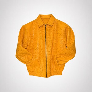 Può includere: Una giacca giallo brillante caratterizzata da una superficie testurizzata con piccoli punti scuri regolarmente distanziati su tutta la superficie. La giacca presenta un colletto standard, una chiusura centrale con zip, maniche lunghe e un orlo elasticizzato in vita.