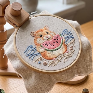 May include: A wooden embroidery hoop stand holds a circular frame with a linen fabric featuring an embroidered hamster eating a slice of watermelon on a sandy beach with blue waves. Three skeins of Airo matte cotton thread, scissors, and a thimble.