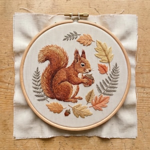 May include: A circular wooden embroidery hoop containing a piece of off-white fabric with a hand-stitched design of a reddish-brown squirrel holding an acorn. The squirrel is surrounded by embroidered autumn leaves and fern fronds in shades of orange, yellow, and grey.