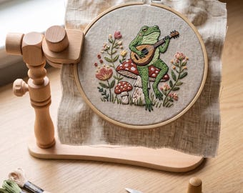 May include: An embroidery hoop mounted on a wooden stand, featuring a green frog playing a lute while sitting on a red and white mushroom, surrounded by embroidered flowers. Nearby are three skeins of embroidery floss and metal embroidery scissors.