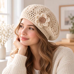 May include: A cream-colored crochet beret featuring an open-knit texture and a prominent, layered flower appliqué on the side. The flower has light cream petals surrounding a darker, brownish-tan center, adding detail to the hat.