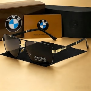Könnte beinhalten: Schwarz-goldene Sonnenbrille mit Doppelsteg-Design und BMW-Logo auf den Bügeln. Die Gläser sind dunkel und mit einem Aufkleber mit der Aufschrift Polarized versehen. Das Set enthält eine braune Box, eine goldene Karte mit BMW-Logo und ein schwarzes Tuch.
