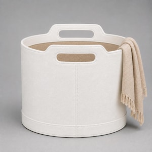 Pode incluir: Um cesto de armazenamento redondo em branco sujo com duas pegas integradas e costuras visíveis nas laterais. Uma manta têxtil bege com franjas está drapeada sobre o lado direito do cesto, contrastando com o exterior liso e claro.