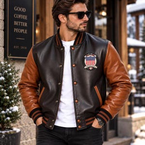 May include: A varsity jacket featuring a dark brown leather body and tan leather sleeves. The jacket has a white t-shirt underneath, a front button closure, and a circular patch on the chest displaying a shield with stars and stripes.