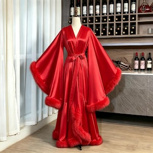 May include: A floor-length red satin robe featuring wide, flowing sleeves and a matching tie-waist belt. The garment is accented with thick, fluffy red faux fur trim along the sleeve cuffs and the entire front opening, extending to the hem.